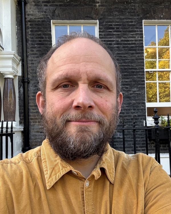 A man with a beard and a brown shirt stands in front of a brick building.
