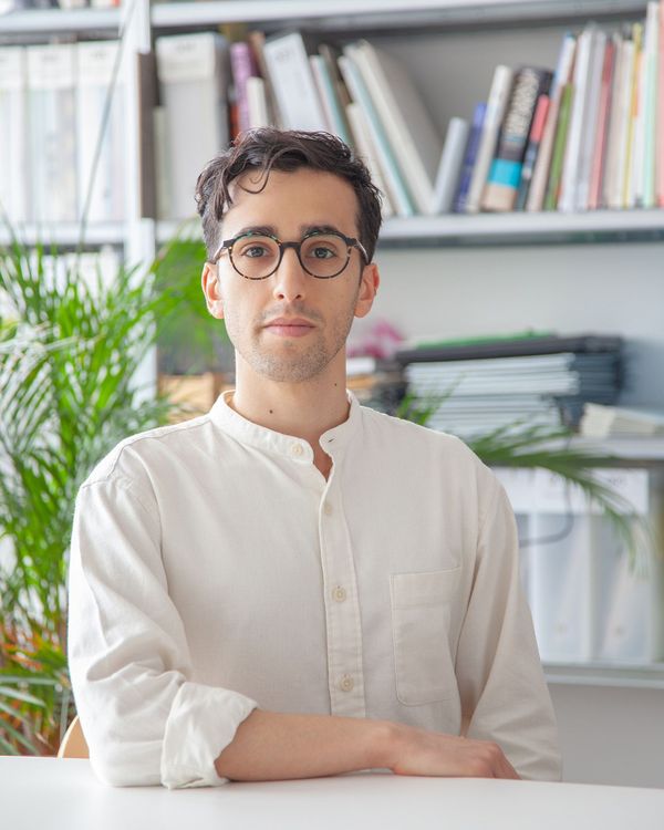 A man with glasses and a white collared shirt sits at a desk in front of a bookshelf.