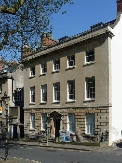 A tall, narrow, light-colored historic building with many windows stands on a sunny city street.