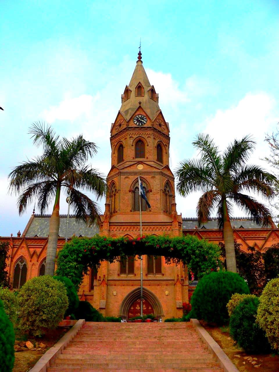 A tall, ornate clock tower rises above a historic building surrounded by palm trees and manicured gardens.