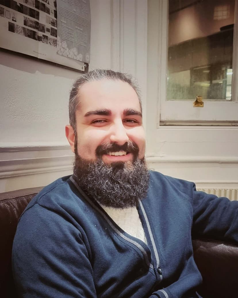 Man with a beard smiling at the camera while sitting indoors.