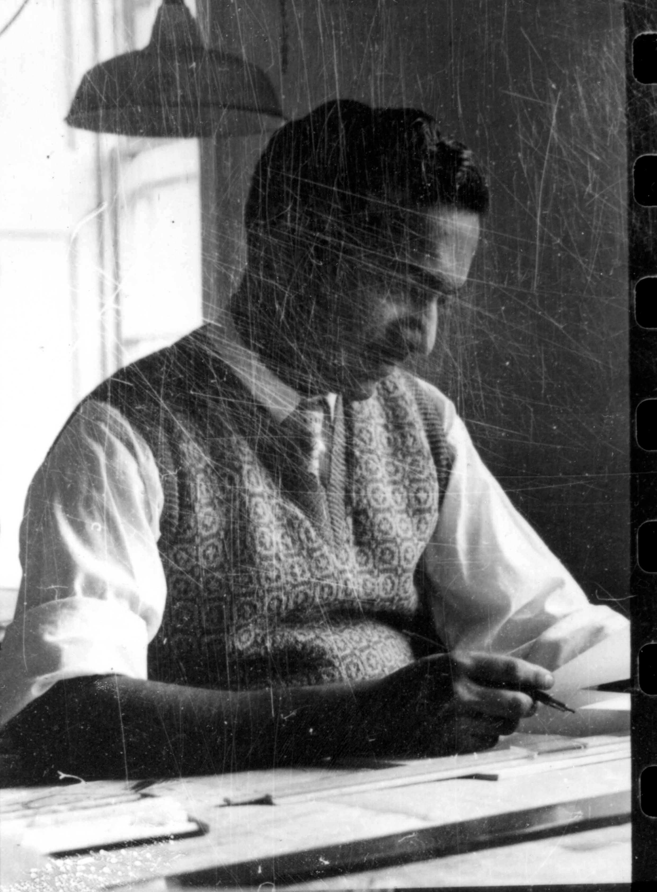Man in a patterned vest and white shirt drawing at a desk under a lamp.