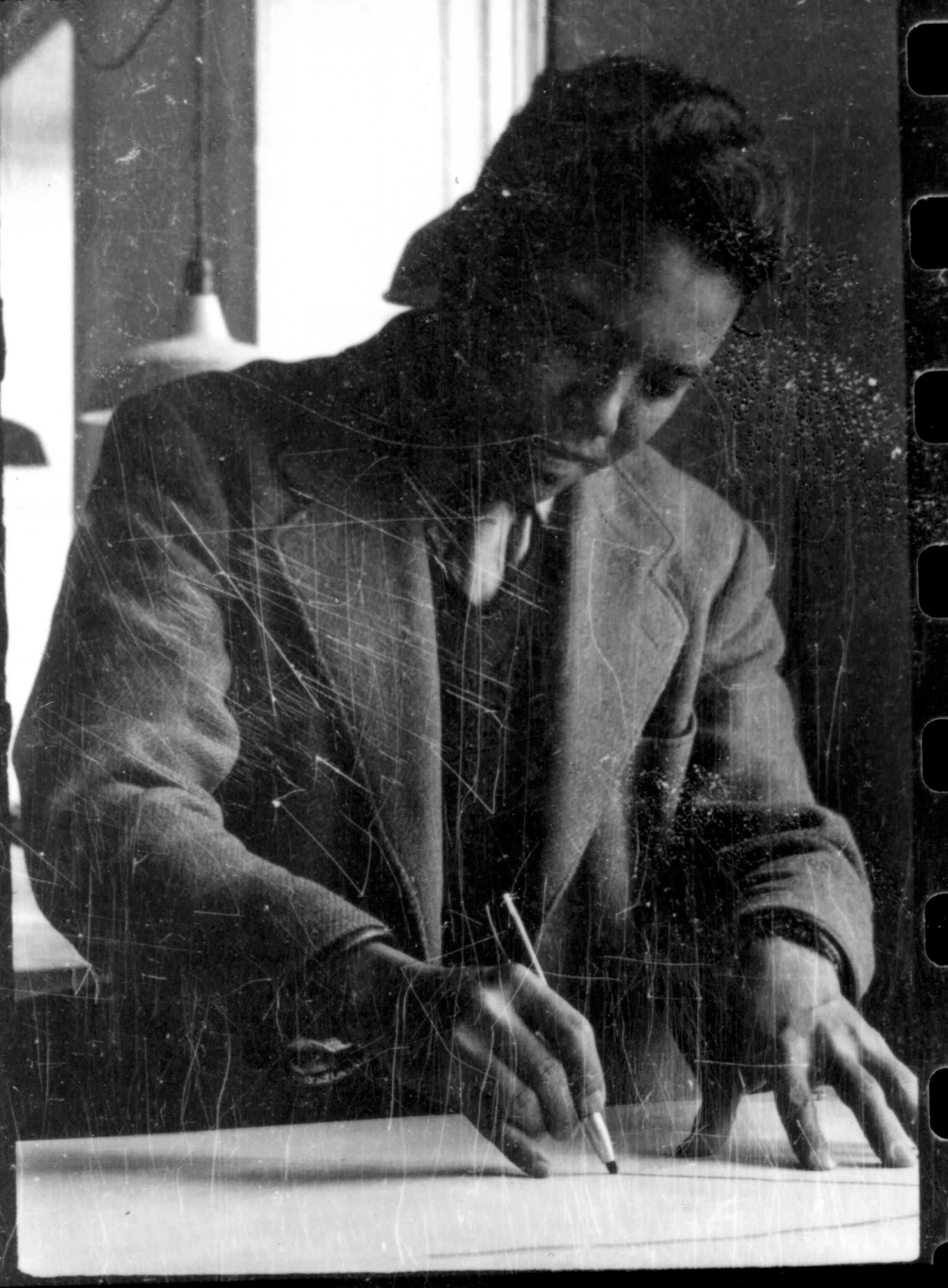 Man in a suit, tie, and cap diligently writing or drawing at a desk.