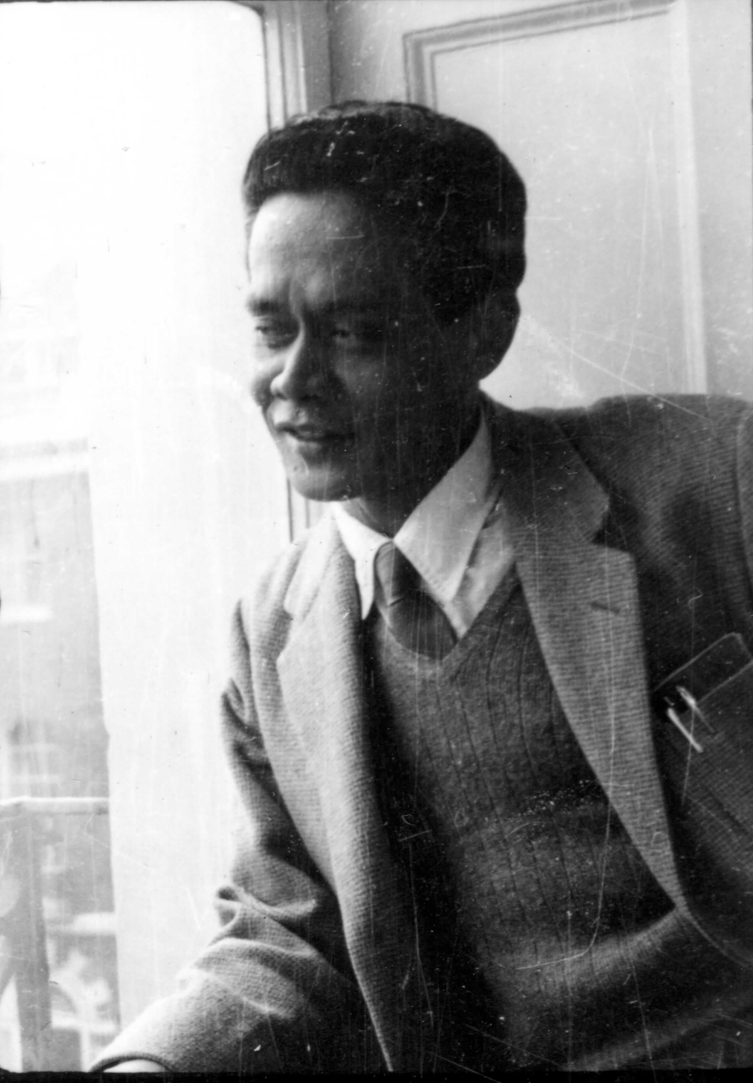 Man in a suit and tie sitting by a window, looking out with a slight smile.