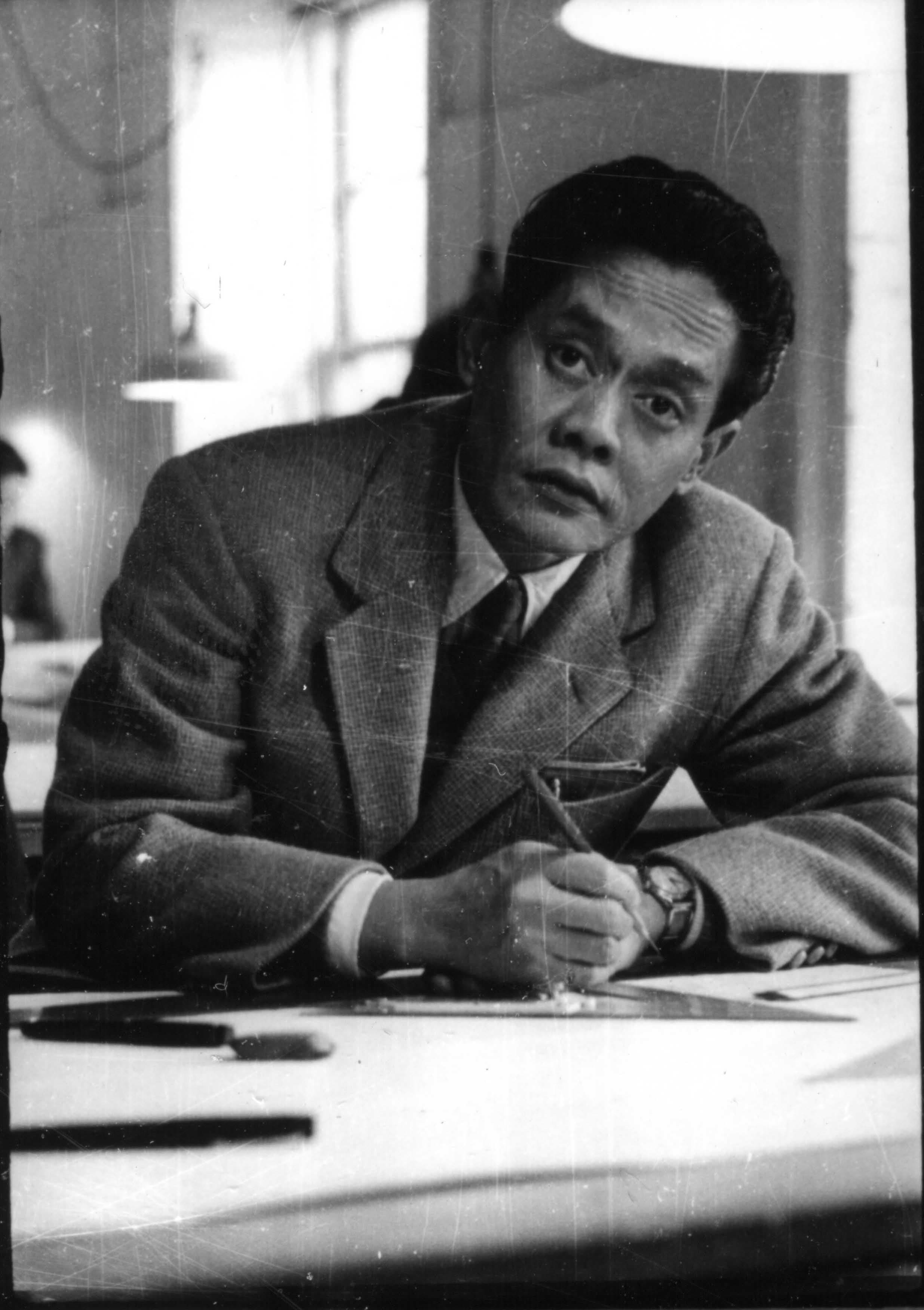 Man in a light suit and tie writing at a desk, looking toward the viewer.