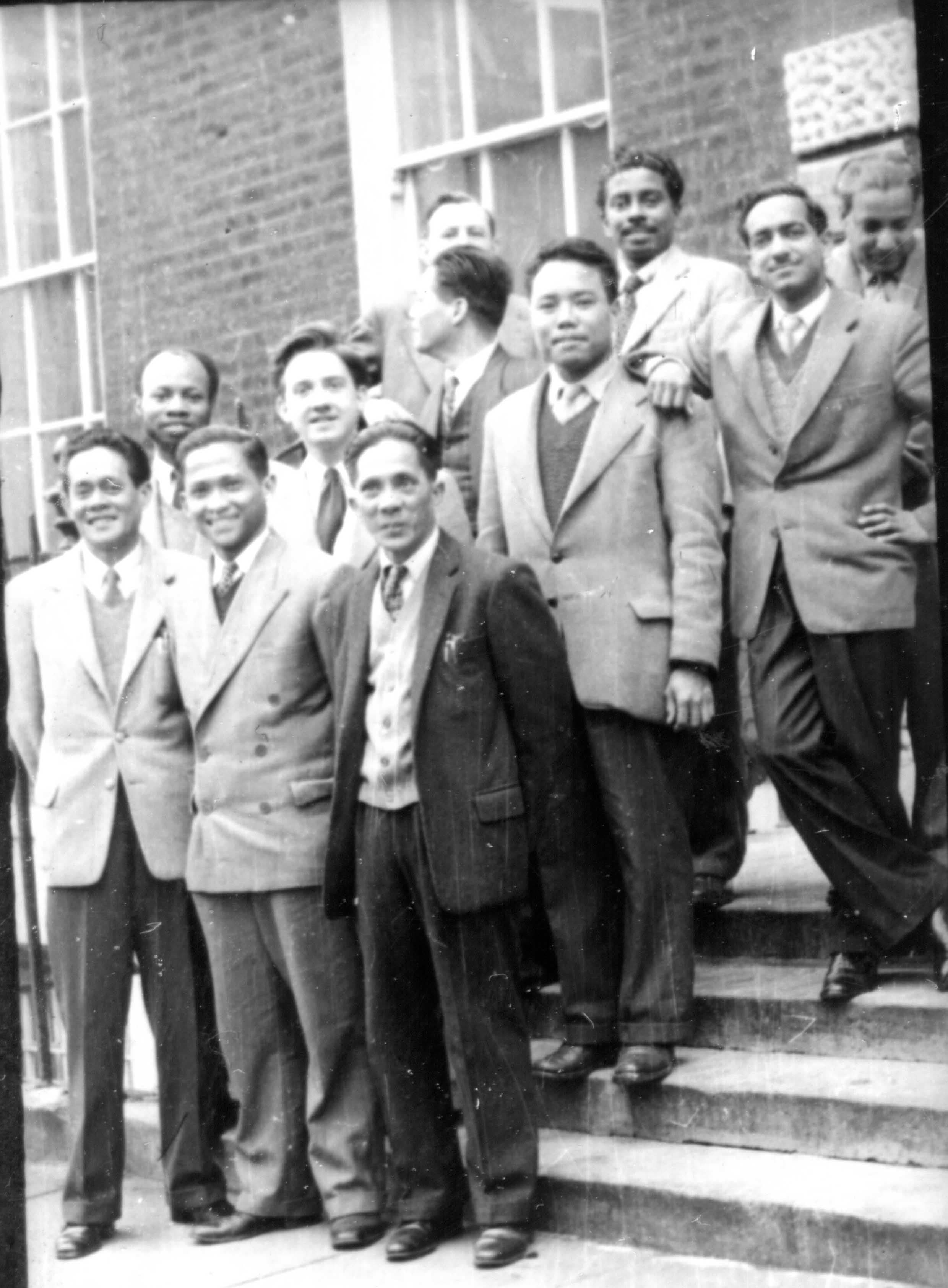 Group of ten men in suits and ties posing on the steps of a brick building.
